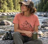 A woman wearing a coral heathered short-sleeve T-shirt with a black line drawing of a bird and the words 'Finch Please' in a natural outdoor setting.